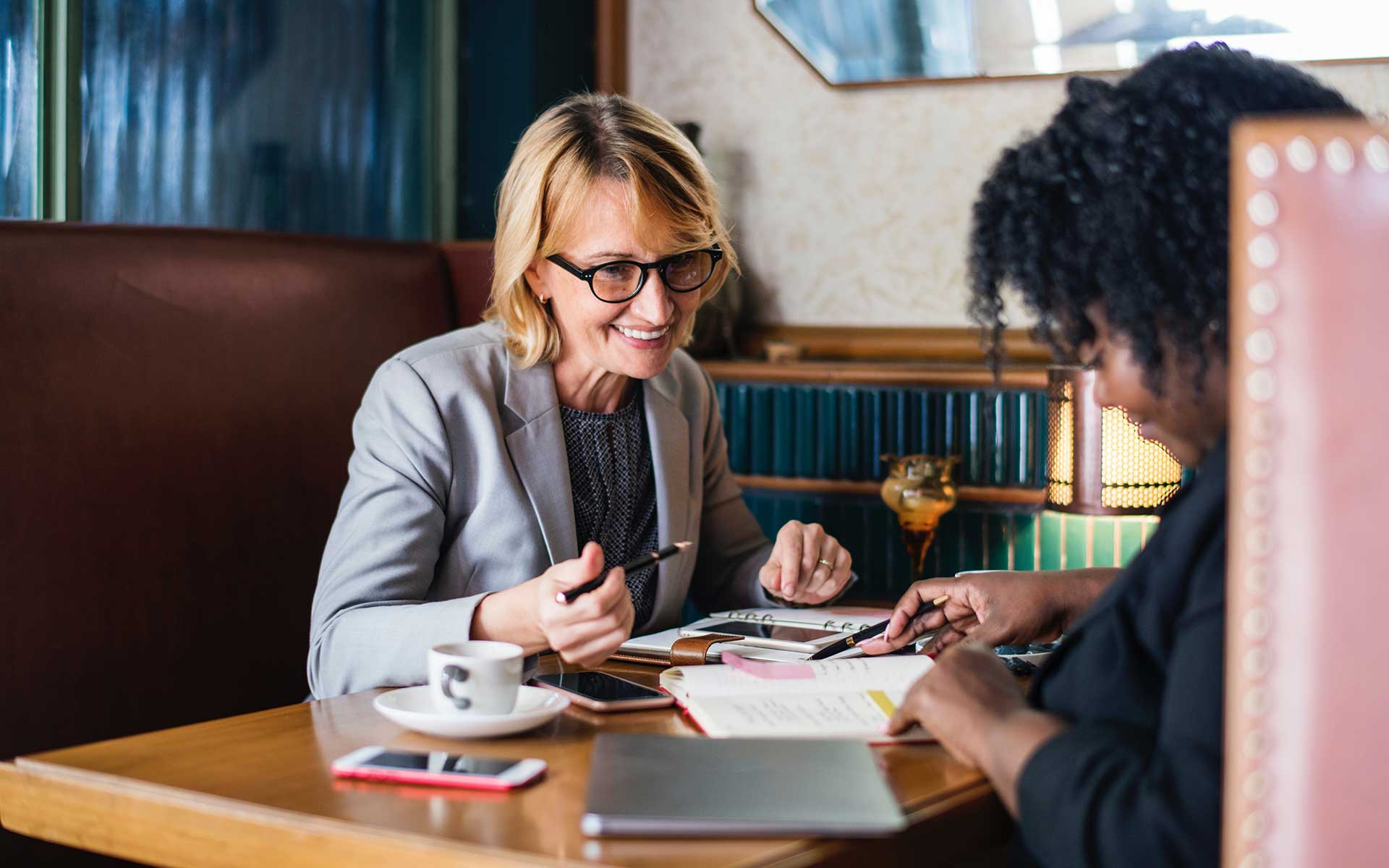 Two women sitting at a table with papers and coffee.